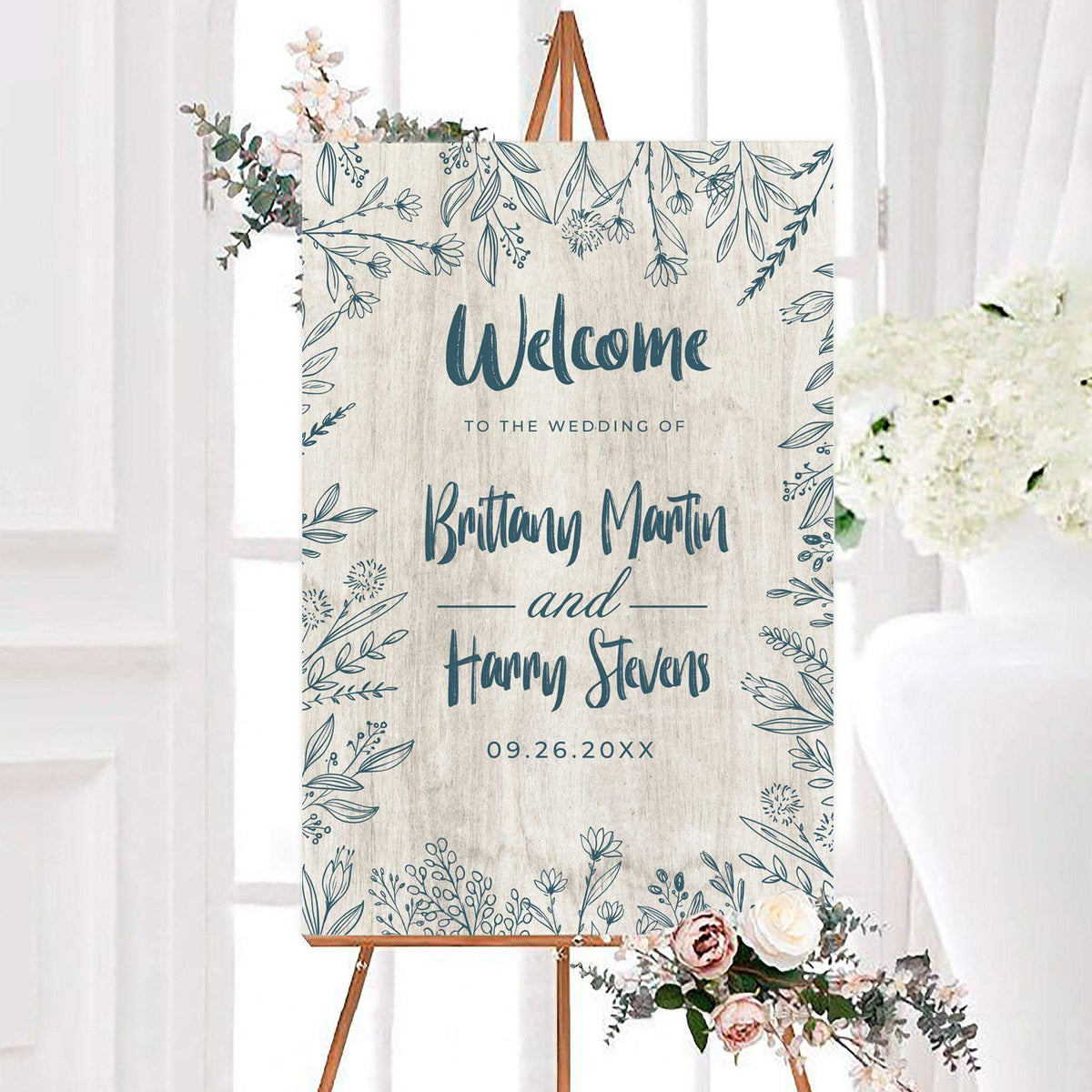 Rustic blue floral frame wedding welcome sign featuring a textured background surrounded by hand-drawn botanical flowers and leafy branches. Personalized script names and a wedding date are displayed prominently in the center.