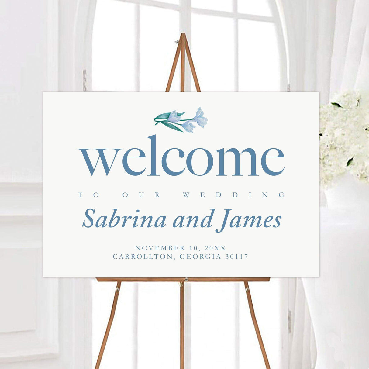 Minimalist blue lily wedding welcome sign featuring soft blue typography and a small watercolor floral accent with green leaves. The design includes personalized couple names, wedding date, and event details on a clean white background.