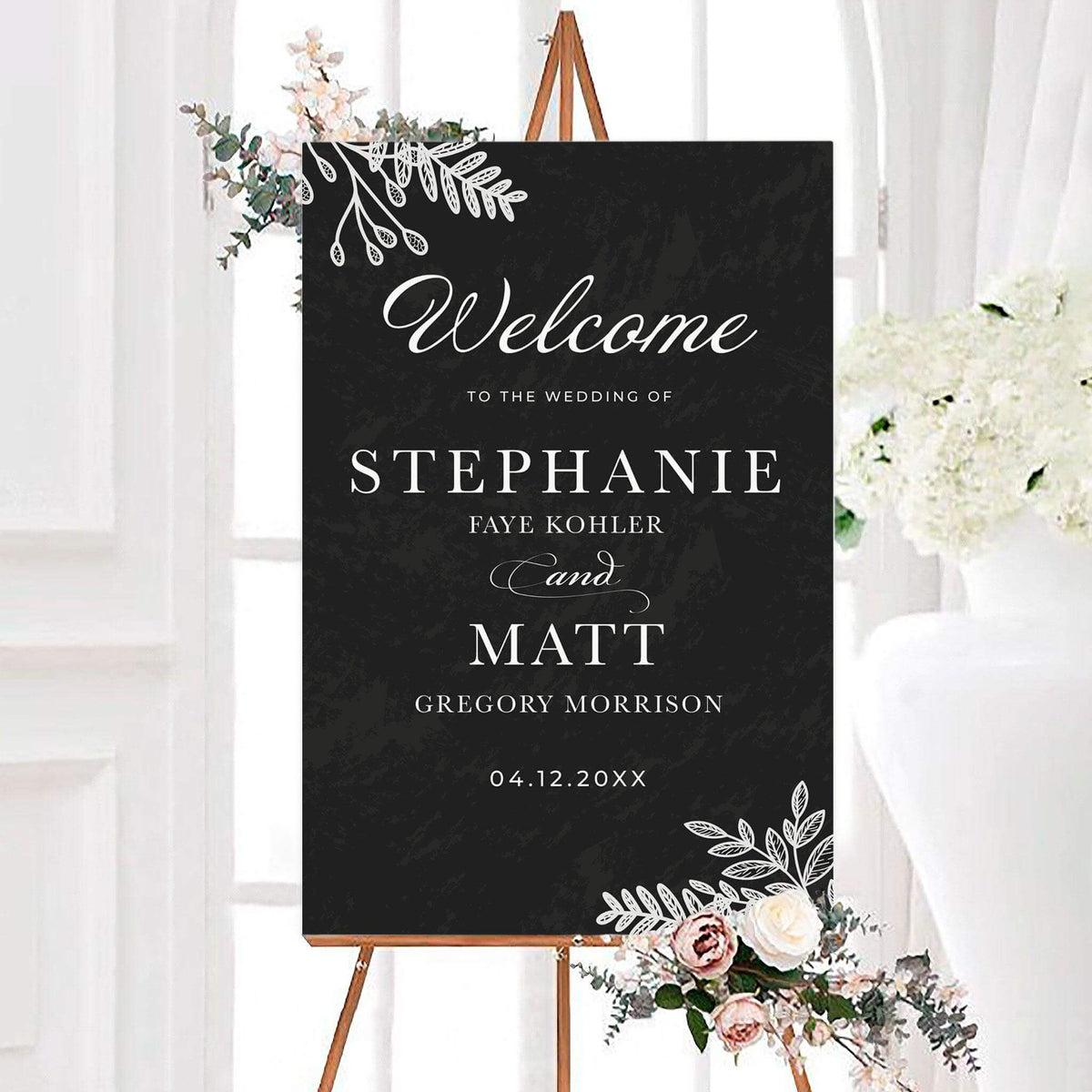 Personalized chalk dusted leaves wedding welcome sign featuring delicate white botanical leaf illustrations, elegant script lettering, and classic serif typography. The charcoal botanical wedding sign includes customizable couple's names and wedding date with decorative leafy corner accents.