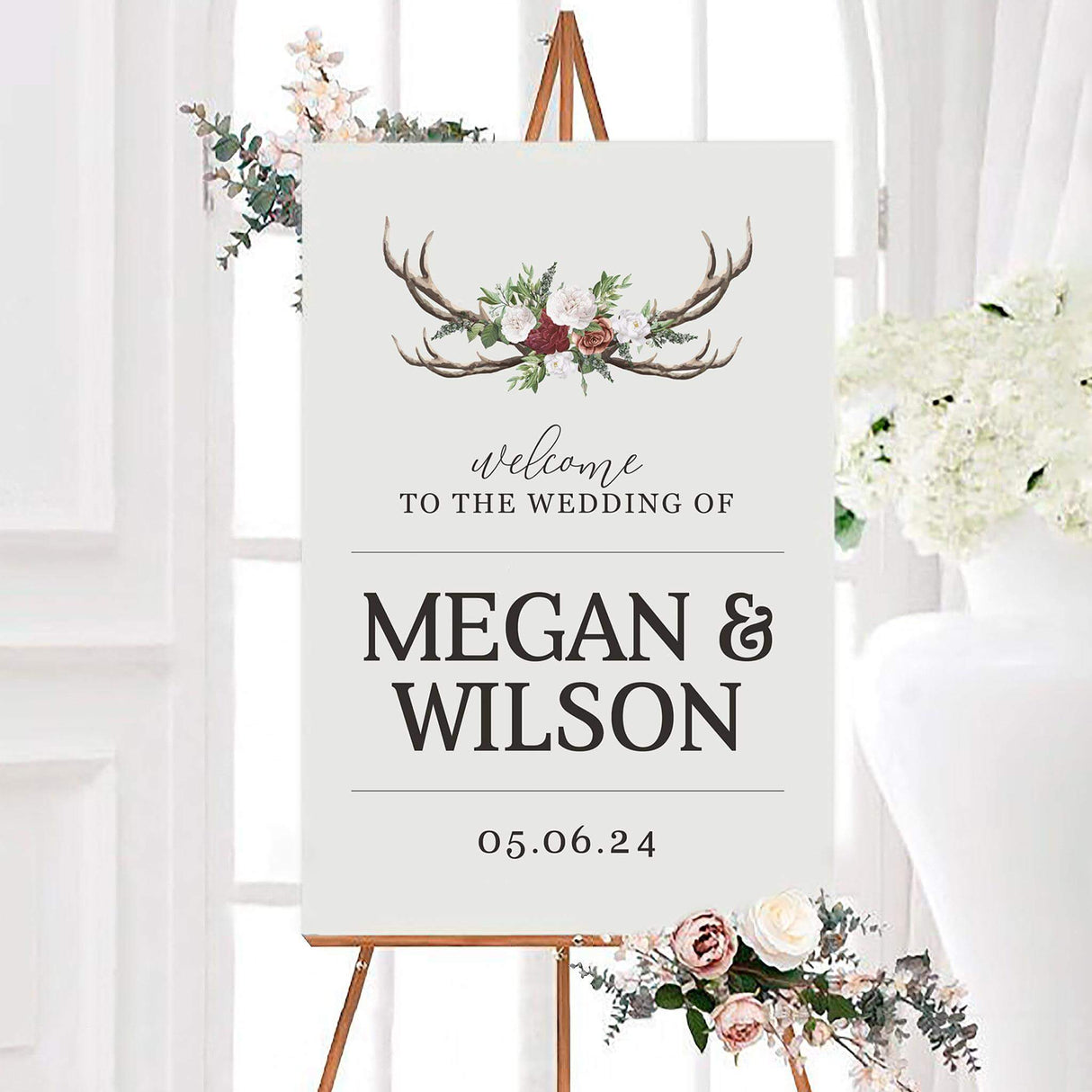Rustic deer horn wedding welcome sign featuring watercolor antlers decorated with burgundy, blush, and white flowers with greenery. Personalized names and a wedding date appear in bold black typography on a light background.