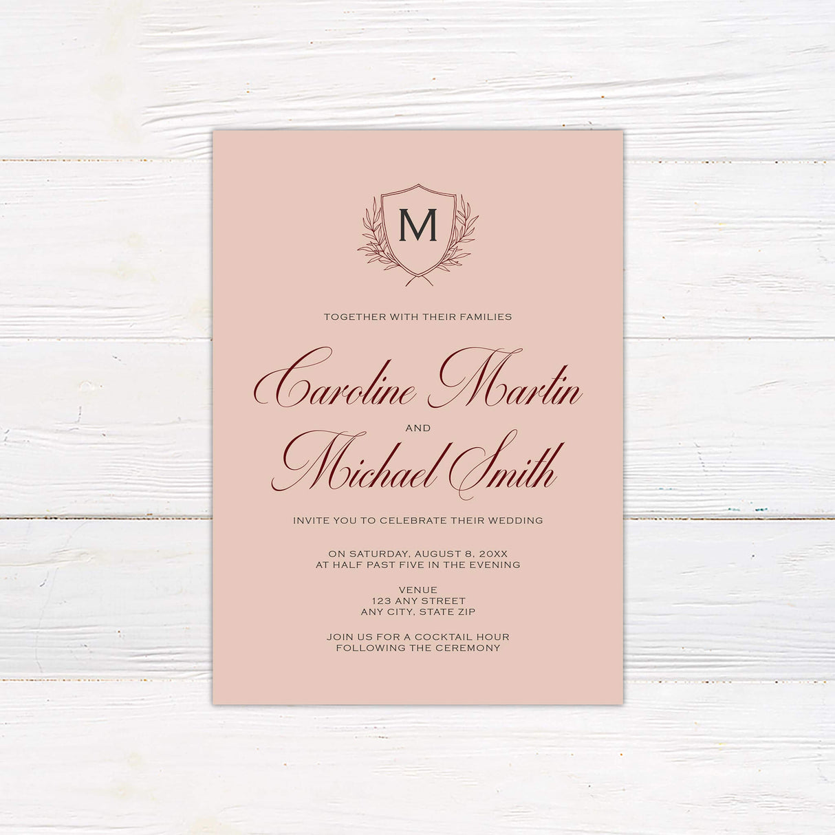 A wedding invitation with a blush background features an elegant monogram crest at the top, accented by delicate laurel details. The text is arranged in a mix of flowing script and classic serif fonts, creating a timeless and sophisticated look. Event details, including the date, time, and venue, are displayed in a structured and refined layout. This design exudes elegance and charm, perfect for a formal, classic, or vintage-inspired wedding celebration.