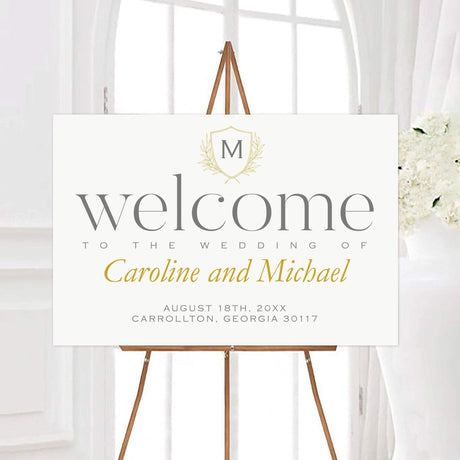 Elegant wedding welcome sign with monogram crest and gold laurel, featuring names and date in calligraphy on ivory background.