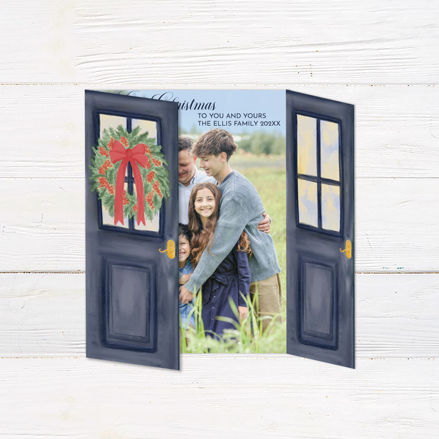 Personalized Doorway to Christmas gatefold card featuring navy blue doors decorated with festive evergreen wreaths, red bows, and elegant gold Merry Christmas lettering. The doors open to reveal a family photograph with a customizable holiday greeting and family name.