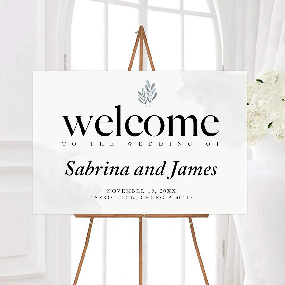 Elegant leaves wedding welcome sign featuring classic black typography and a delicate botanical leaf accent. The design displays the couple’s names, wedding date, and location on a clean light background.