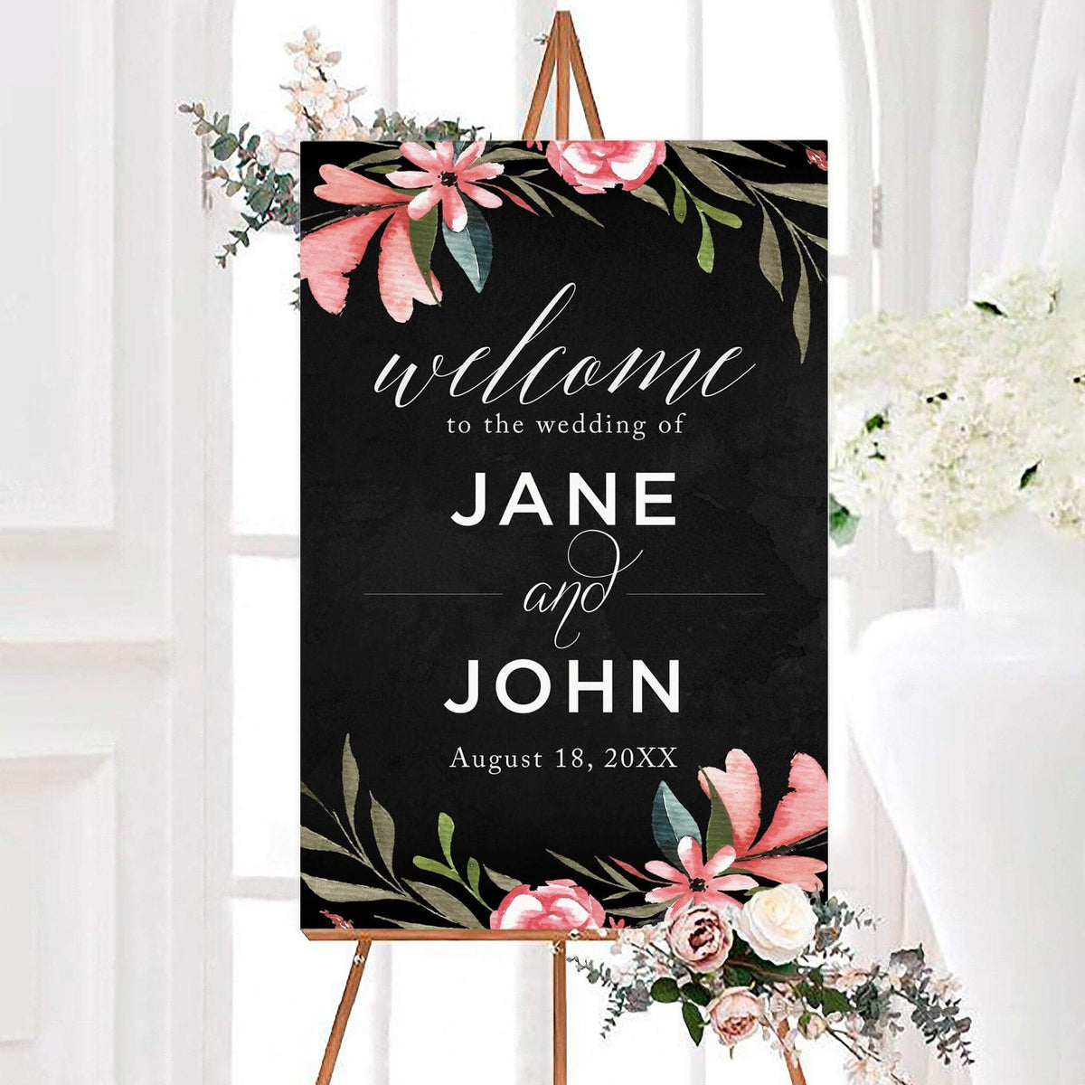 Floral suede wedding welcome sign with pink watercolor flowers and greenery on a dark background. Personalized white lettering displays the couple’s names and wedding date.