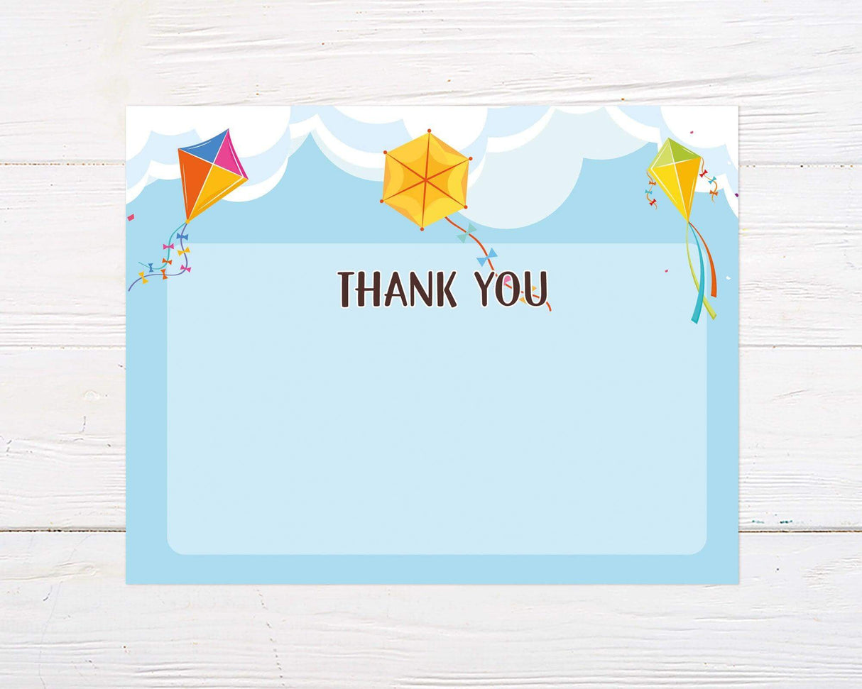A colorful thank you card features three bright kites flying against a soft blue sky with fluffy white clouds. The words "Thank You" are written in playful, bold lettering at the center of the design.