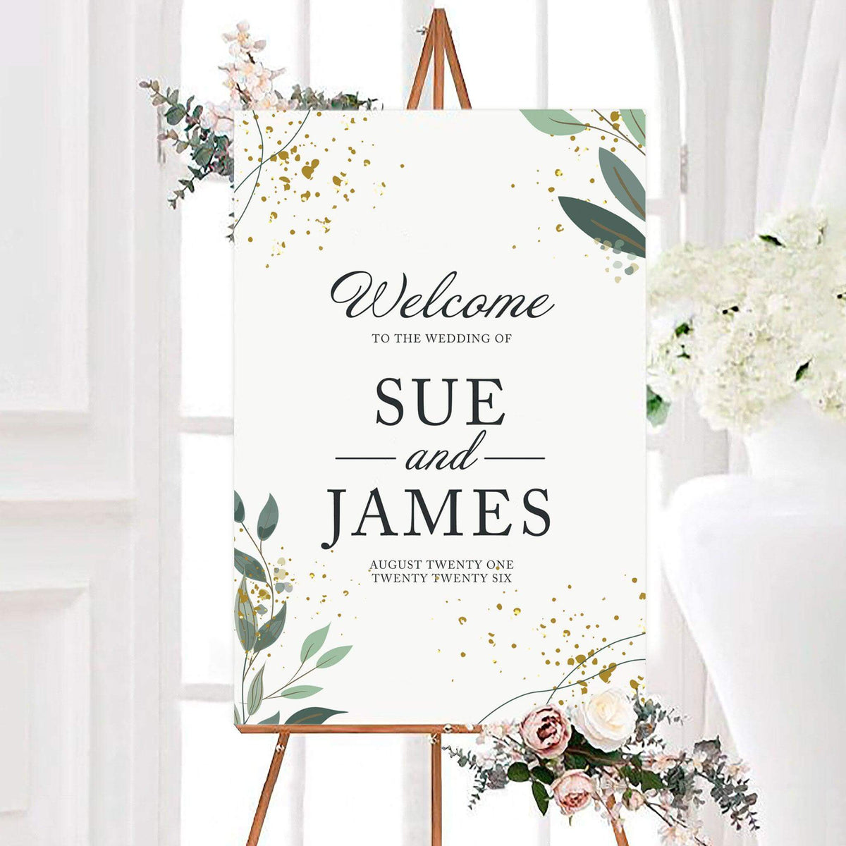 Personalized gold flake greenery wedding welcome sign featuring leafy greenery and scattered metallic-inspired decorative speckles. The couple’s names and wedding date are displayed in elegant serif and script typography.
