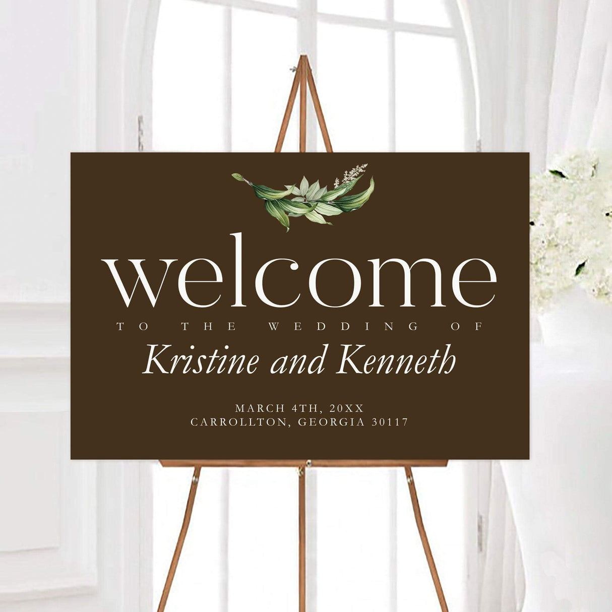 Elegant gold frame foliage wedding welcome sign featuring a rich brown background, white typography, and a small watercolor greenery accent. The personalized design includes the couple’s names, wedding date, and event location.