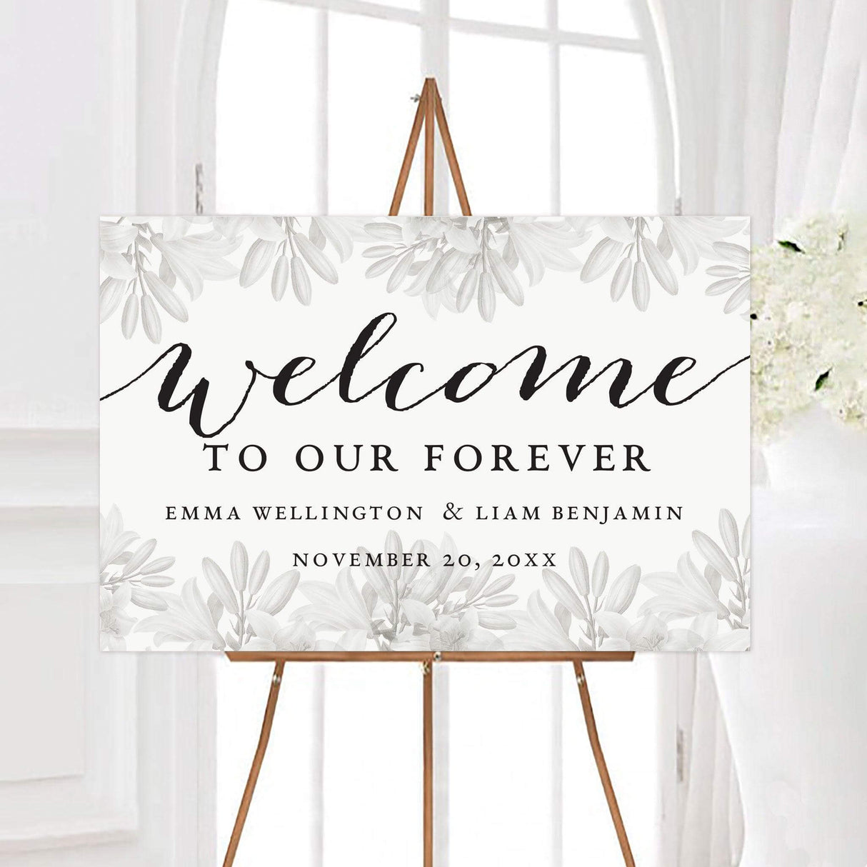 Elegant grayscale floral wedding welcome sign featuring soft white and gray lilies framing black calligraphy and serif typography. The design displays the couple’s names and wedding date on a clean white background.