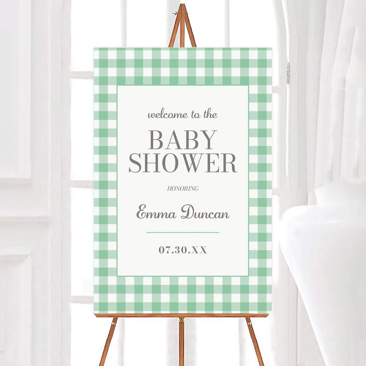 Baby shower welcome sign with a green and white gingham border. The sign features elegant serif text reading "Welcome to the Baby Shower" and includes the honoree's name and event date. Displayed on an easel in a bright setting.