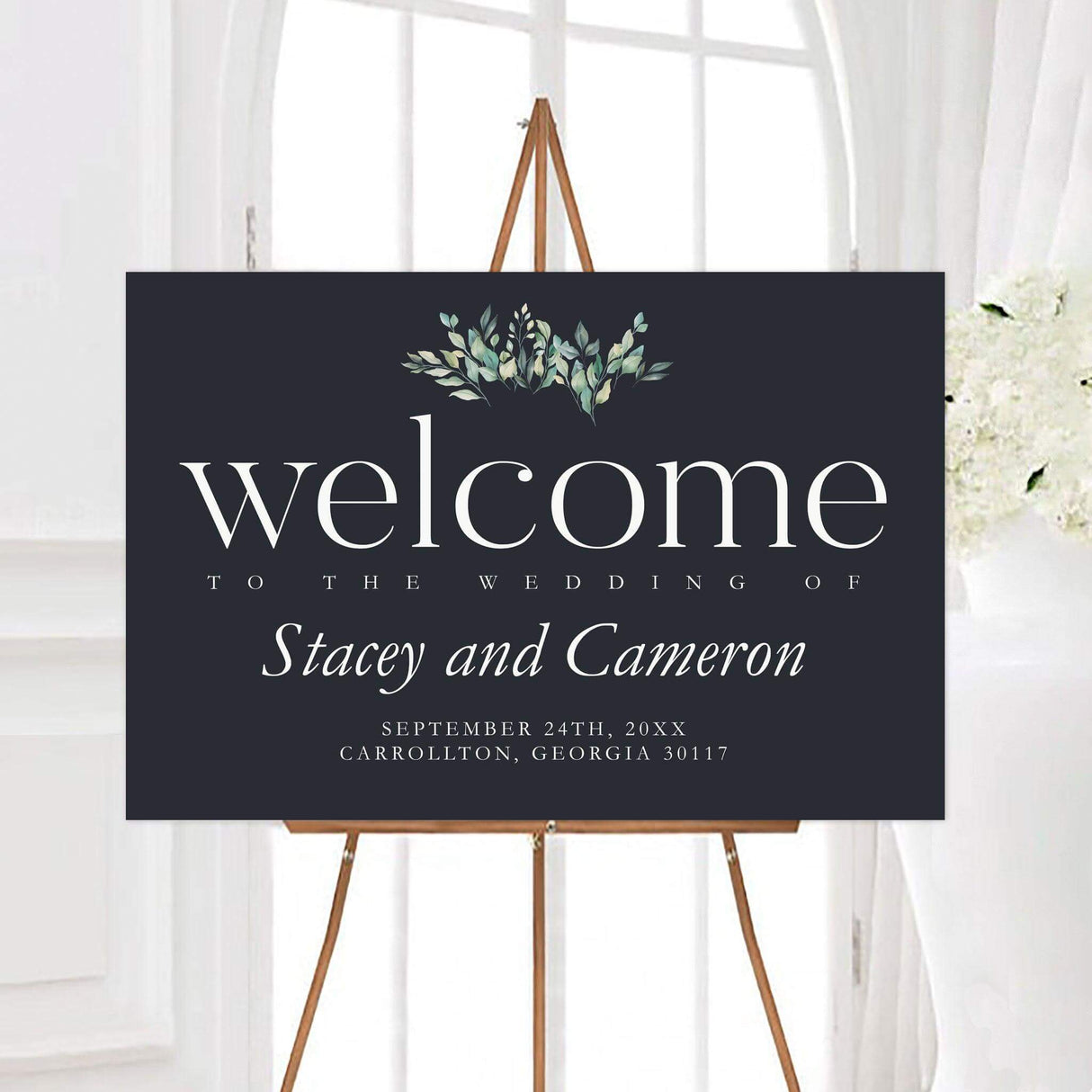 Elegant greenery abundance wedding welcome sign with a dark charcoal background, white typography, and soft watercolor greenery at the top. The sign features personalized couple names, wedding date, and optional location.