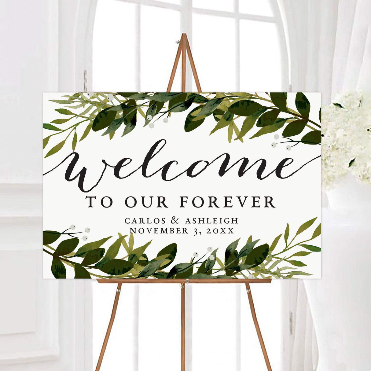 Personalized greenery arch wedding welcome sign framed with watercolor green leaves, branches, and delicate white berries. Black script and serif lettering display a welcome message, couple’s names, and wedding date.