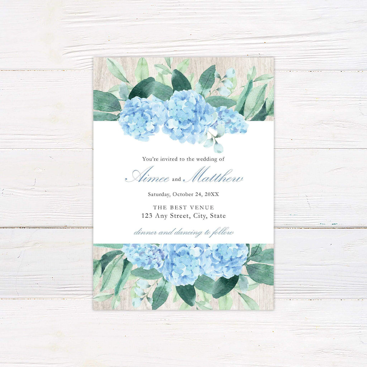Wedding invitation with watercolor blue hydrangeas and greenery on a wood-textured background with elegant script and serif fonts.