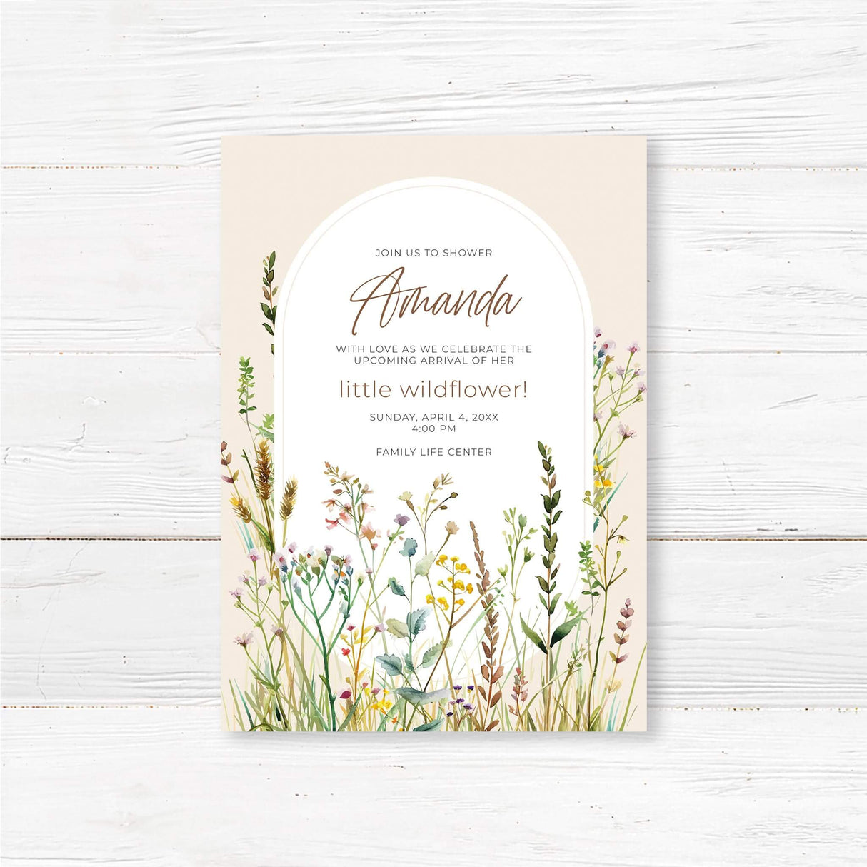 A printed baby shower invitation featuring a soft beige background with a white arched frame in the center containing the event details. The text is in elegant, modern script and serif fonts. The bottom half of the design showcases watercolor-style wildflowers in pastel shades of pink, yellow, green, and purple. The invitation conveys a rustic, nature-inspired theme with a delicate and feminine aesthetic.