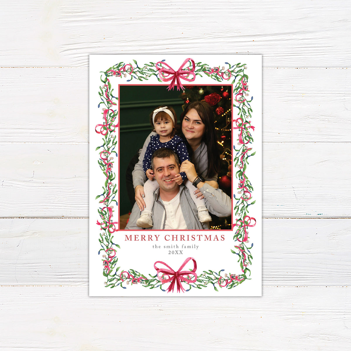 Personalized Christmas photo card featuring a family portrait framed by watercolor mistletoe, greenery, berries, and ribbon bows. The back features a coordinating watercolor bow.