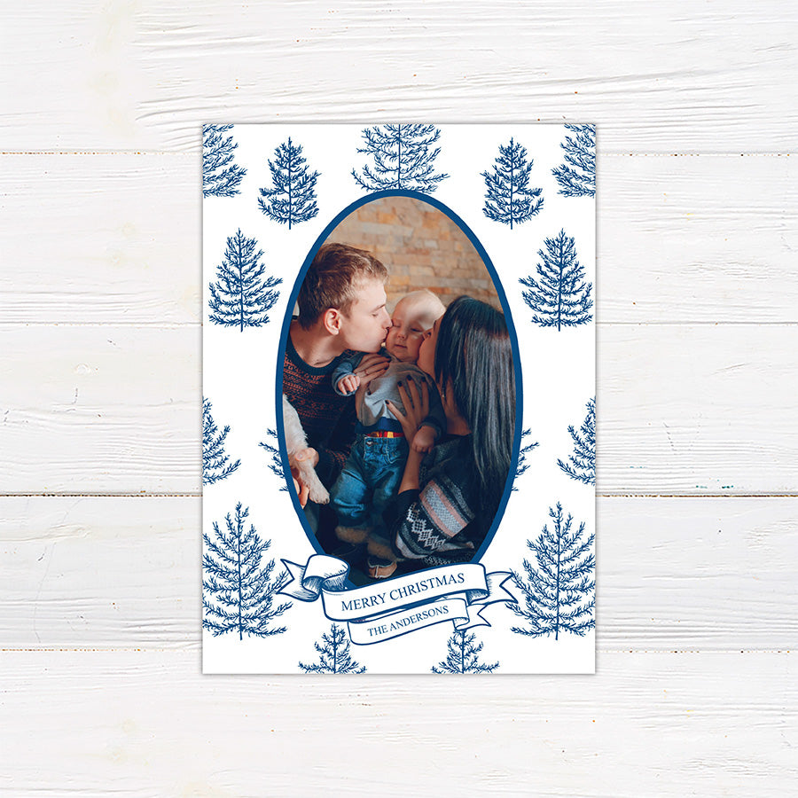 Personalized family Christmas photo card featuring an oval portrait surrounded by hand-drawn evergreen pine trees. A ribbon banner displays a Merry Christmas greeting and custom family name. blue