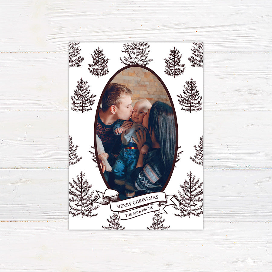 Personalized Winter Pine family Christmas photo card featuring an oval portrait surrounded by hand-drawn evergreen pine trees. A ribbon banner displays a Merry Christmas greeting and custom family name. brown