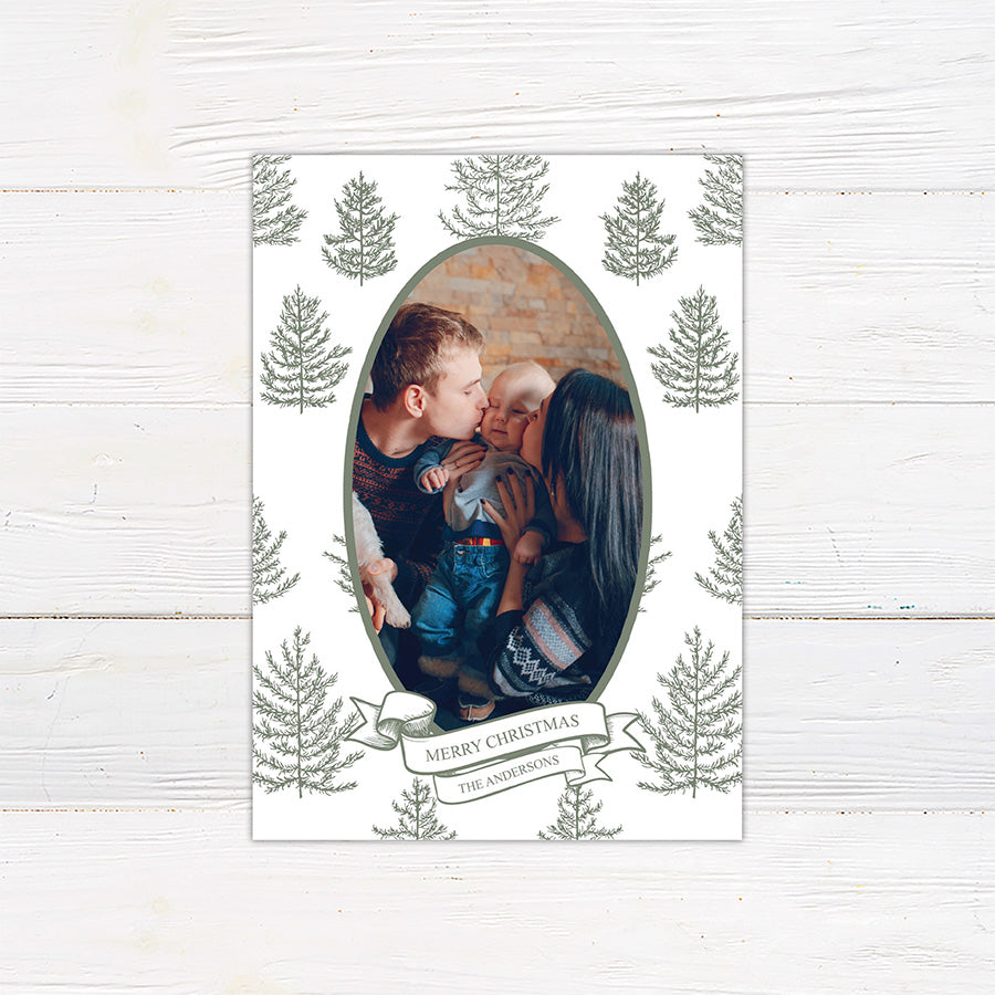 Personalized Winter Pine family Christmas photo card featuring an oval portrait surrounded by hand-drawn evergreen pine trees. A ribbon banner displays a Merry Christmas greeting and custom family name. green