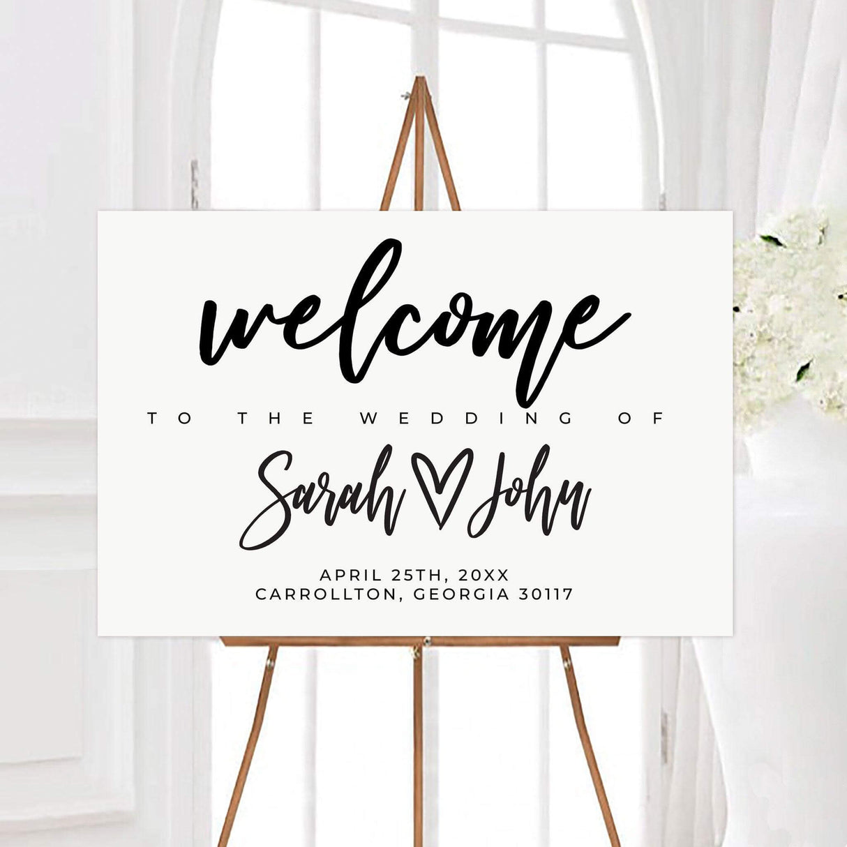 Modern heart wedding welcome sign featuring bold script names with a heart between the couple’s names. The minimalist design includes the wedding date and location beneath the personalized lettering.