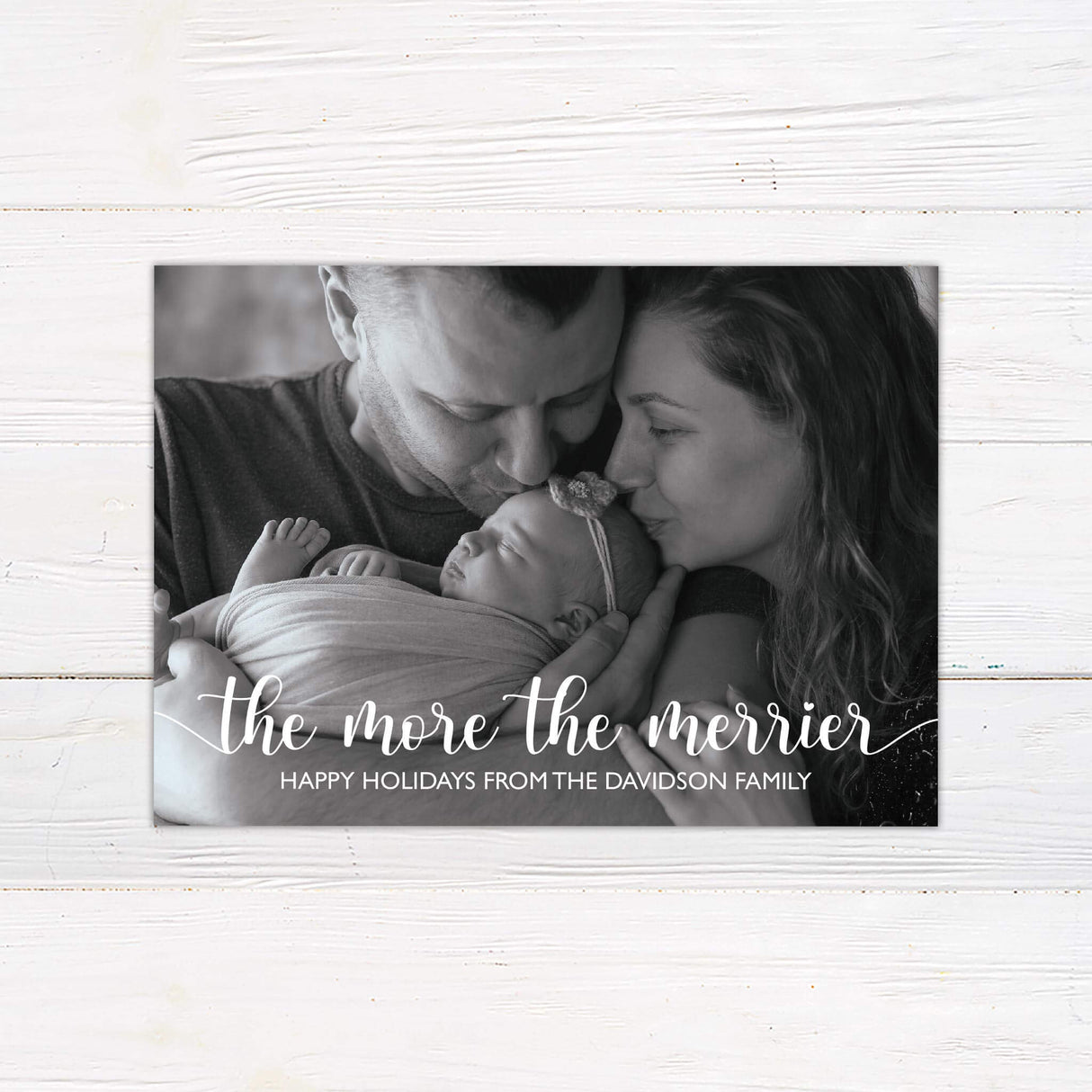 A holiday card design with a family holding their newborn baby and the words “The More the Merrier” displayed in elegant script. The card includes a customizable holiday greeting.