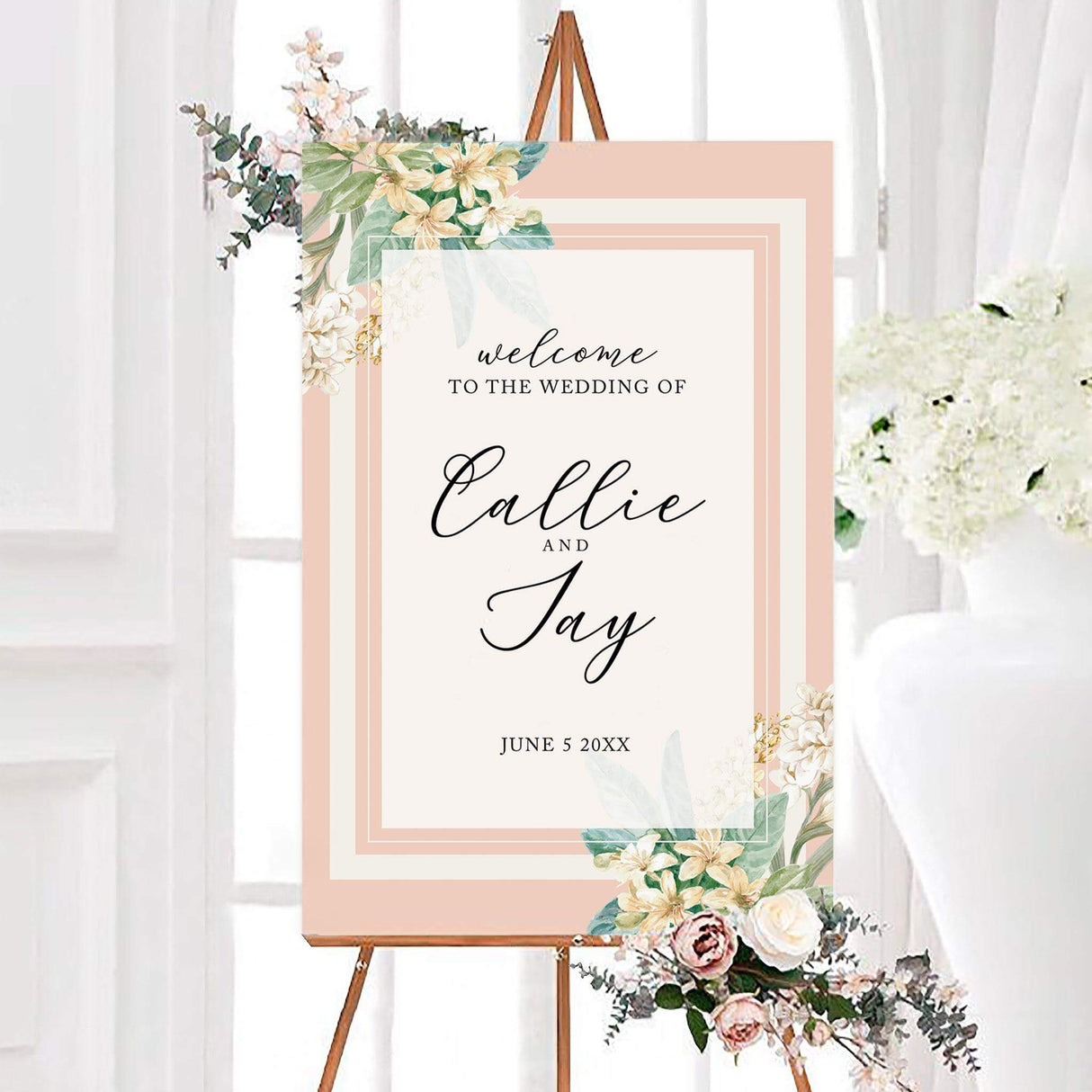 Large wedding welcome sign with white and blush flowers, green leaves, and elegant serif and script text on a cream background.