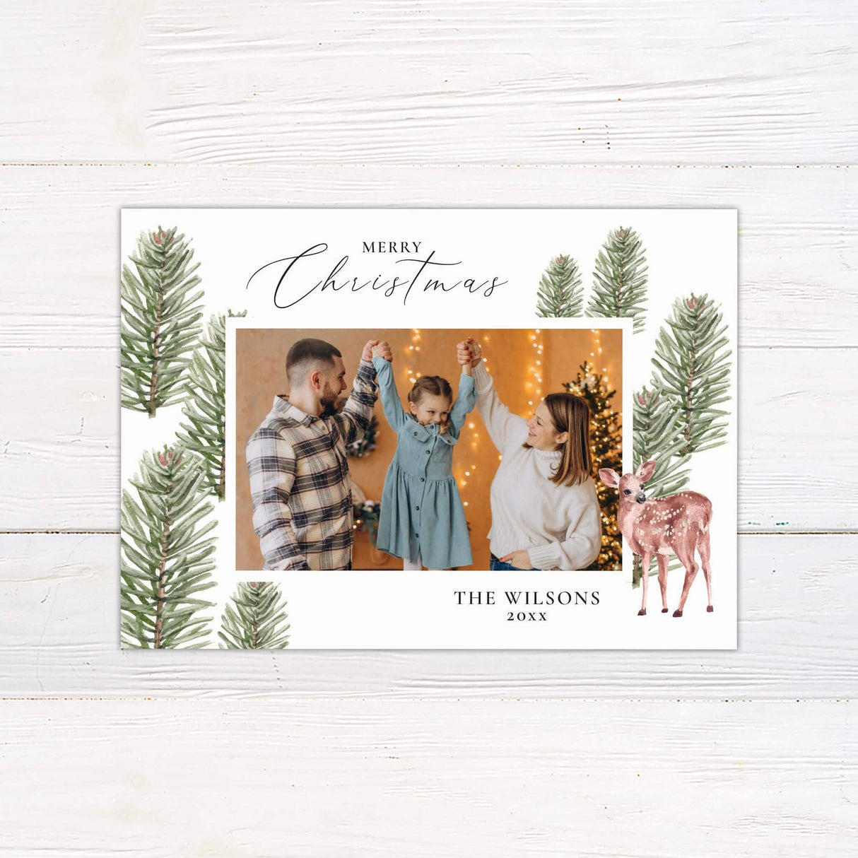 A holiday card design with watercolor pine branches framing a photo and a reindeer illustration on the side. The card layout highlights one large central photo.
