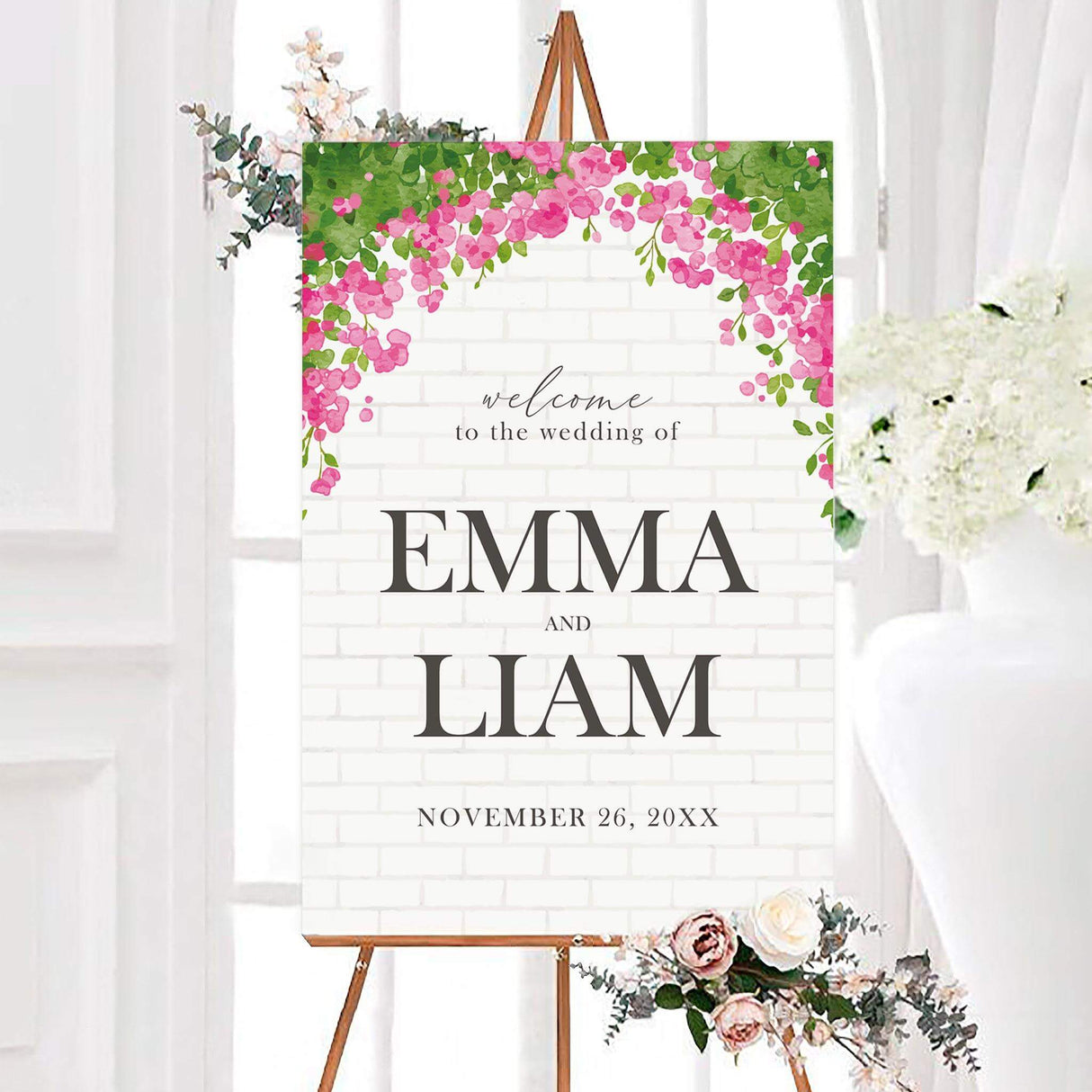 Pink floral arch wedding welcome sign featuring cascading watercolor blossoms and green leaves over a subtle white brick background. Personalized dark gray lettering displays the couple’s names and wedding date.