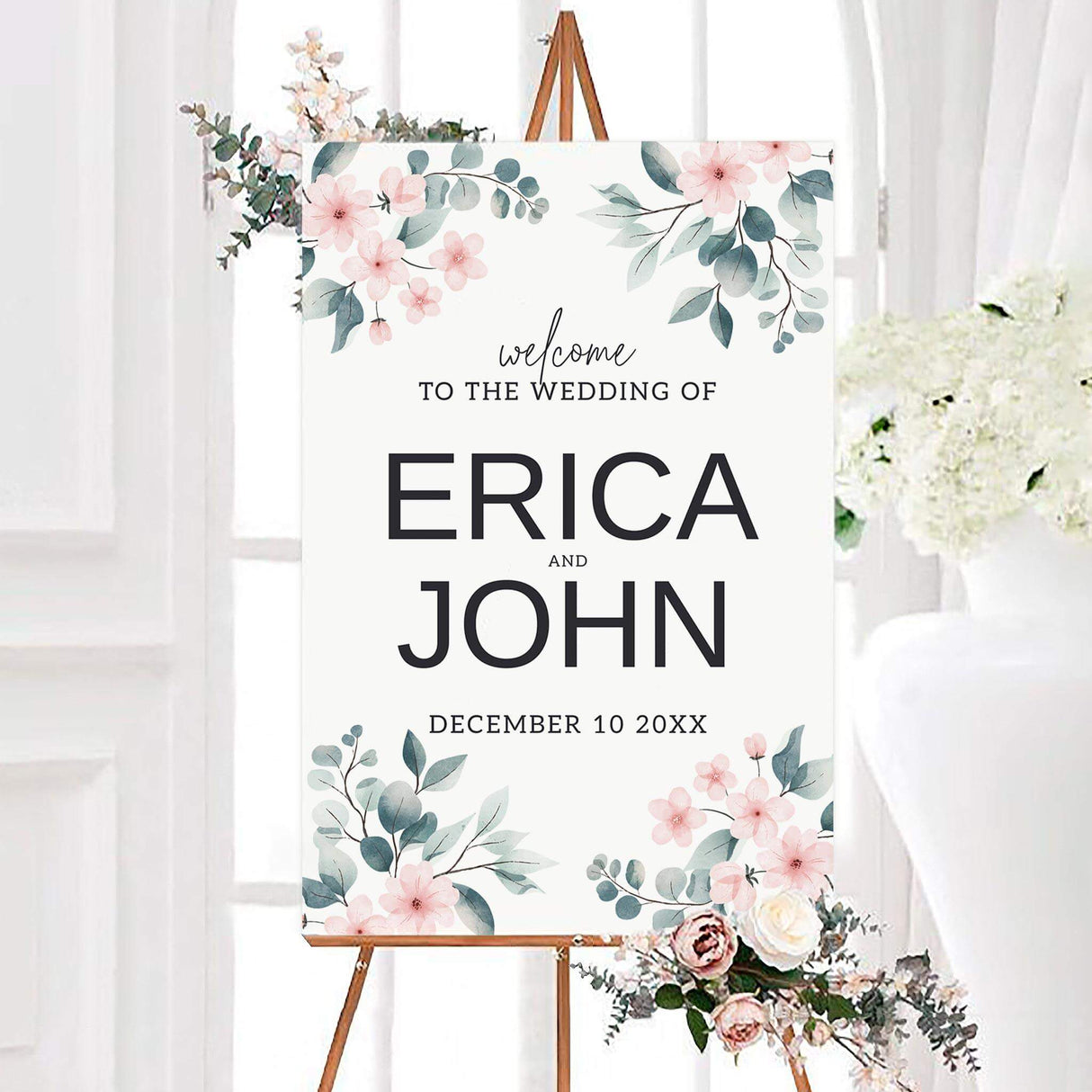 Pink petals wedding welcome sign with watercolor blush flowers, eucalyptus leaves, and bold personalized names. The white botanical design includes a custom wedding date and modern black lettering.