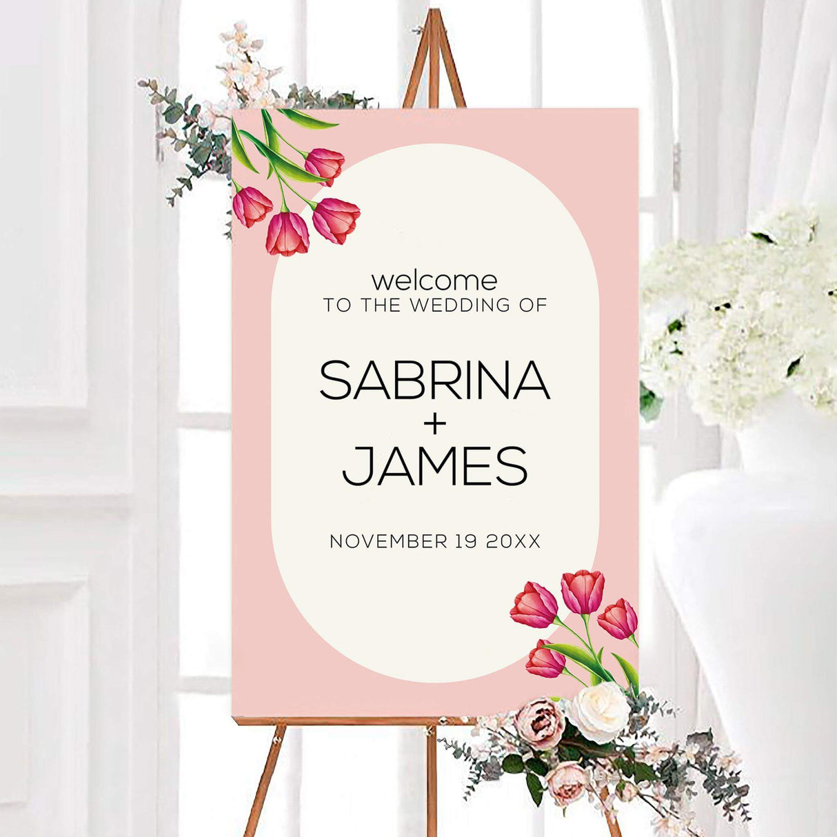 Pink tulip wedding welcome sign with a blush background, ivory arch, and modern black personalized lettering. Bright pink tulips and green leaves decorate the upper and lower corners.