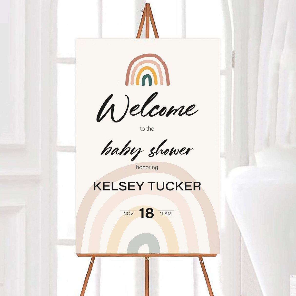Minimalist baby shower welcome sign featuring a neutral color palette with soft earthy tones. The design includes a modern boho-style rainbow at the top and a larger, muted rainbow at the bottom, creating a warm and inviting look. Elegant script and clean typography display the words "Welcome to the baby shower" along with the honoree's name and event details. The sign is printed on high-quality material and displayed on an easel, making it a perfect decorative piece for greeting guests at the entrance of t