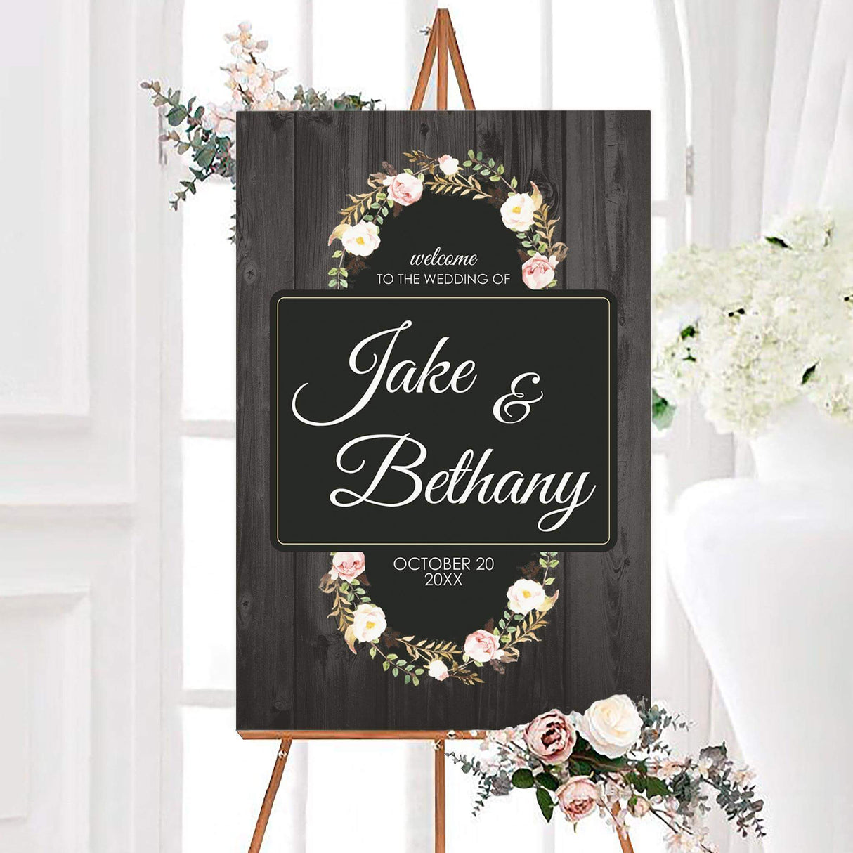 Rustic rose wreath wedding welcome sign with a dark wood background, blush rose wreath, and white script couple names. The personalized floral sign includes an elegant bordered center panel and wedding date.