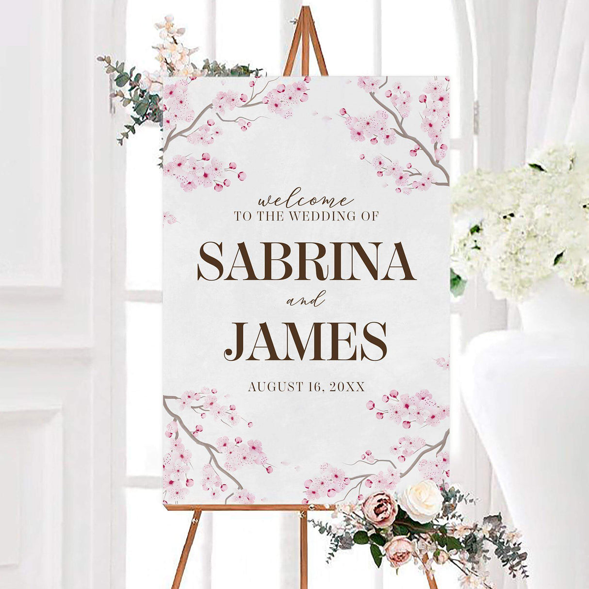 Personalized sakura flowers wedding welcome sign featuring pink sakura cherry blossoms surrounding elegant brown script and serif lettering. The sign displays the couple’s names and wedding date on a soft white background.