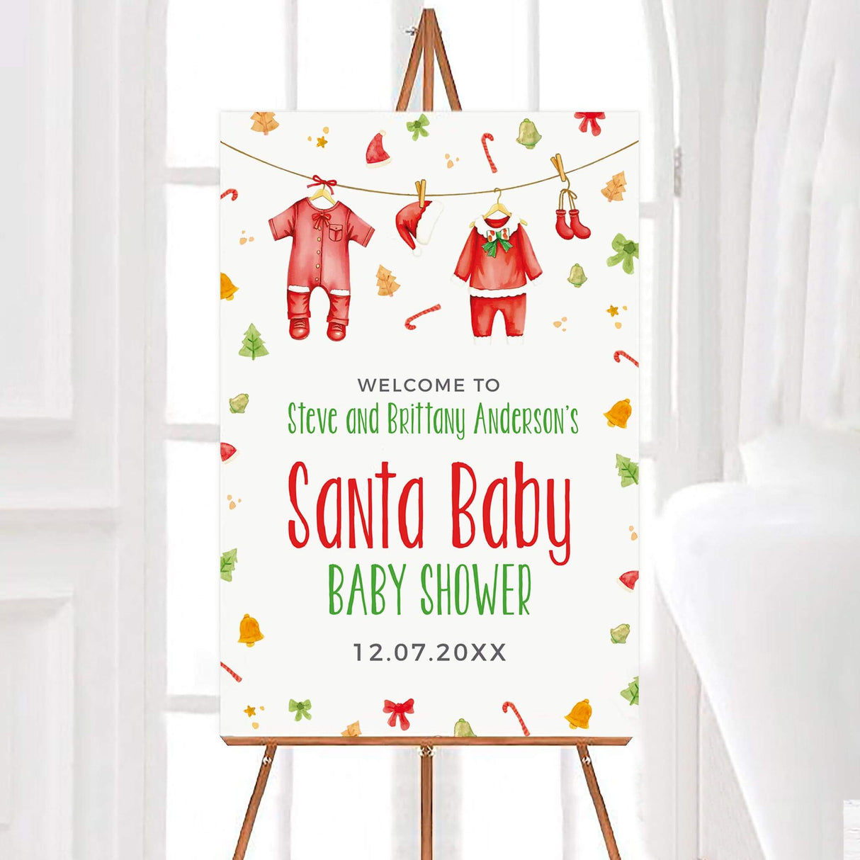 Santa Baby shower invitation with Christmas theme, featuring baby outfits and festive decorations, on an easel with white background.
