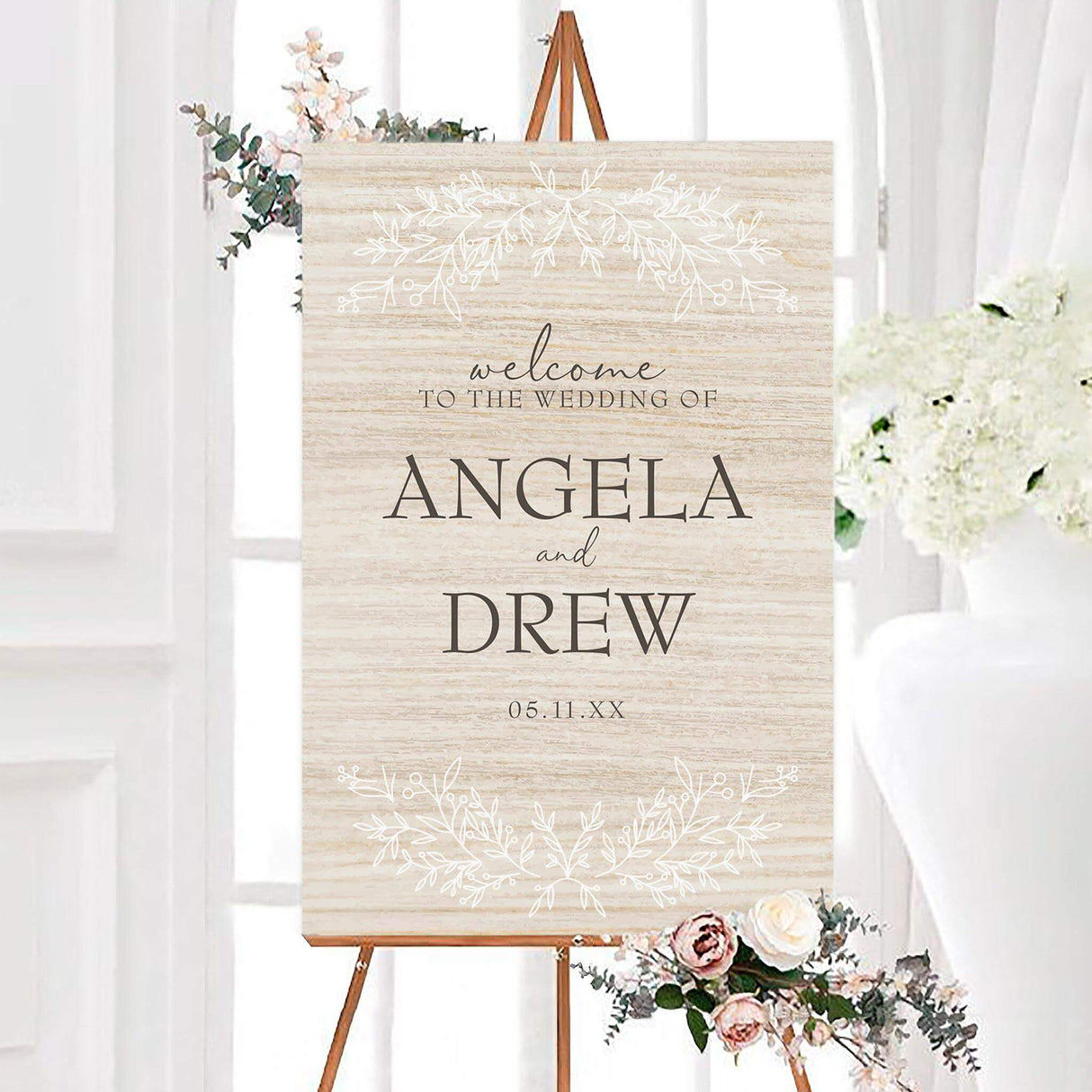 Simple rustic wedding welcome sign with a neutral wood-textured background, personalized couple names, and elegant black typography. White botanical branches and leaves decorate the top and bottom of the sign.