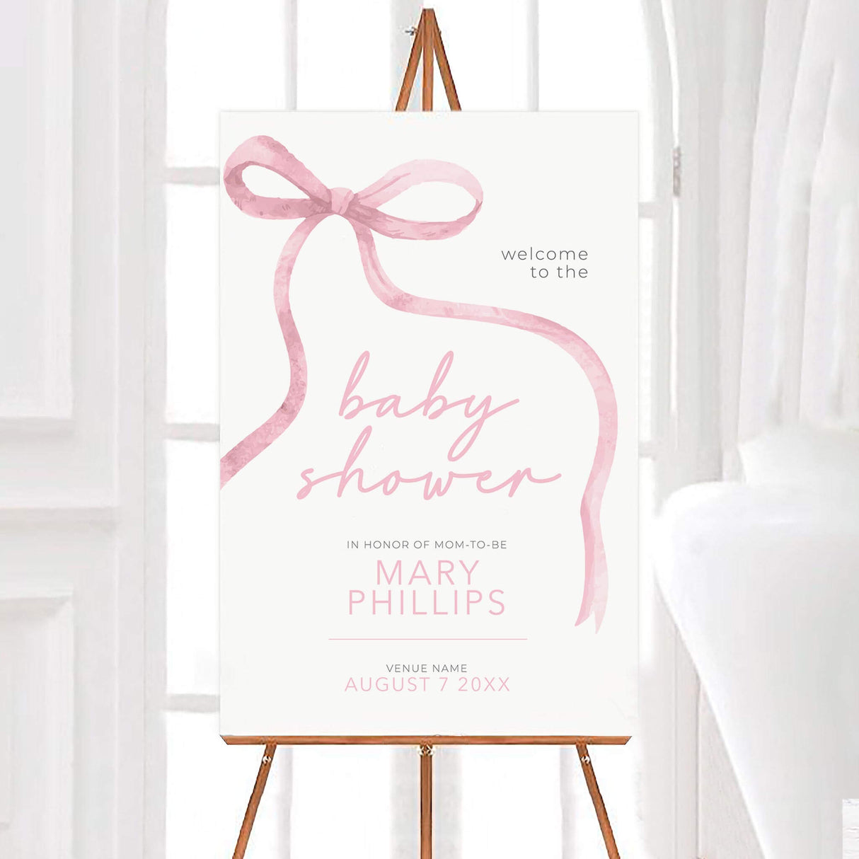 Soft ribbon baby shower invitation with pink watercolor bow and elegant typography displayed on an easel in a bright room.