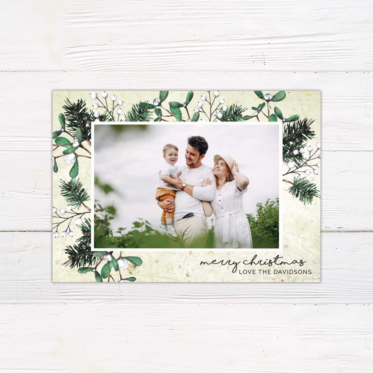 A Christmas card featuring a family photo with pine and mistletoe accents. The design includes a customizable holiday greeting at the bottom.