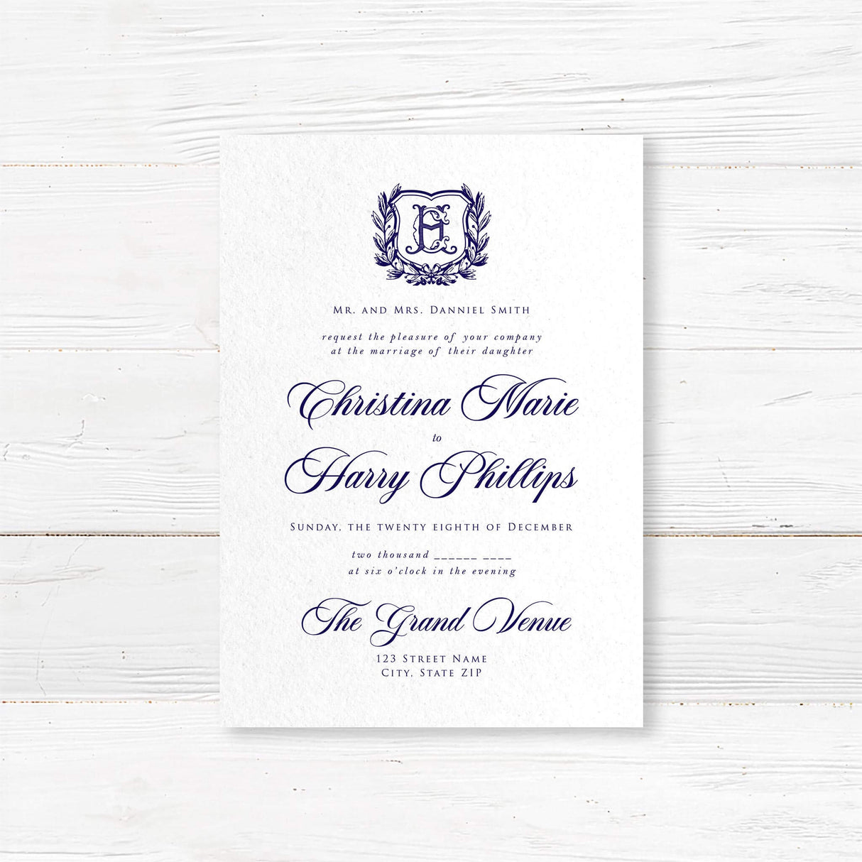 Formal wedding invitation with a vintage-style shield and laurel wreath monogram. Features elegant script for the names and classic serif text for wedding details.