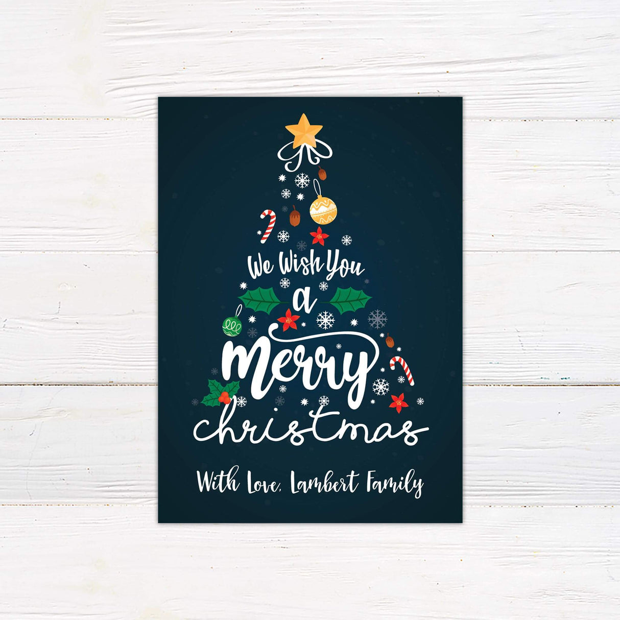 A Christmas card featuring a tree made of ornaments, holly, and snowflakes with the text “We Wish You a Merry Christmas.” The card is customizable with your family name.