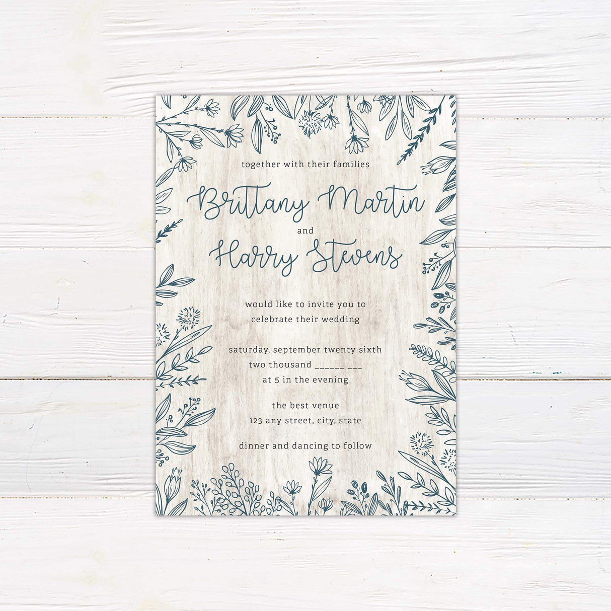 Rustic wedding invitation with hand-drawn blue-gray botanical border on a white wood background and script and serif fonts.