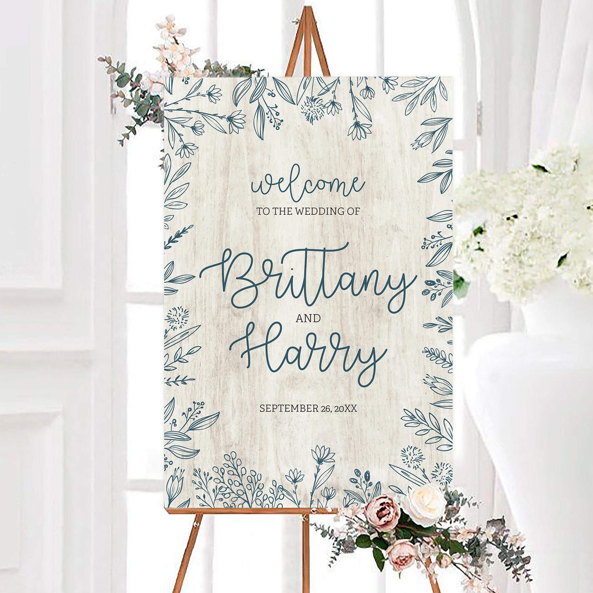 Large welcome sign with rustic white wood backdrop and botanical leaf corner accents.