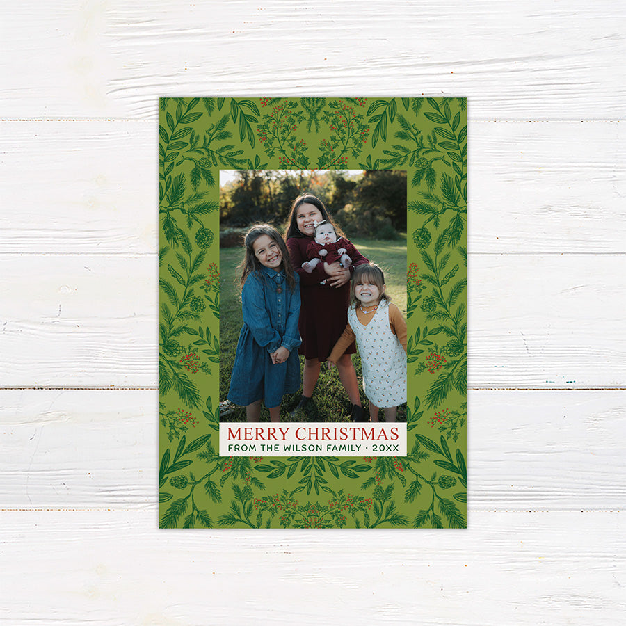 Personalized family Christmas photo card featuring winter greenery, berries, pinecones, and a large family portrait. The coordinating back includes three additional family photos surrounded by botanical holiday artwork.