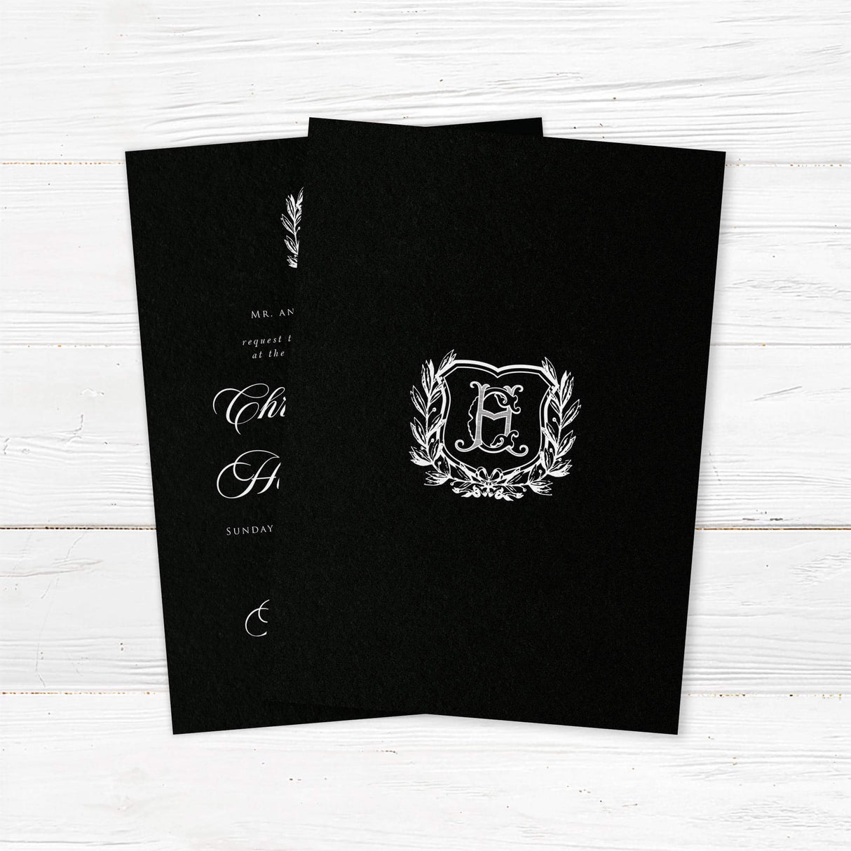 Elegant wedding invitation with black background, vintage-style white shield monogram surrounded by a laurel wreath, white calligraphy and serif fonts, ideal for formal or gothic weddings. - back side