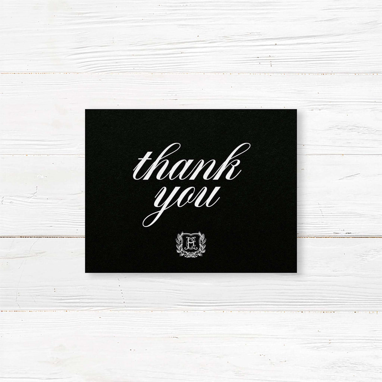 Wedding thank you card with black background, white calligraphy fonts, and an ornate shield monogram encircled by a laurel wreath, matching a formal black wedding suite.