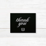 Wedding thank you card with black background, white calligraphy fonts, and an ornate shield monogram encircled by a laurel wreath, matching a formal black wedding suite.