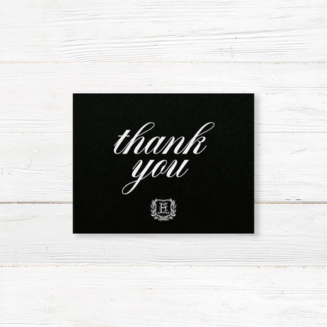 Wedding thank you card with black background, white calligraphy fonts, and an ornate shield monogram encircled by a laurel wreath, matching a formal black wedding suite.