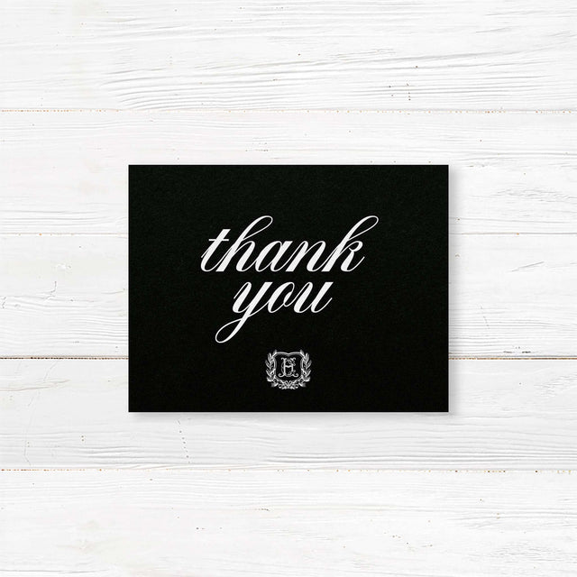 Wedding thank you card with black background, white calligraphy fonts, and an ornate shield monogram encircled by a laurel wreath, matching a formal black wedding suite.