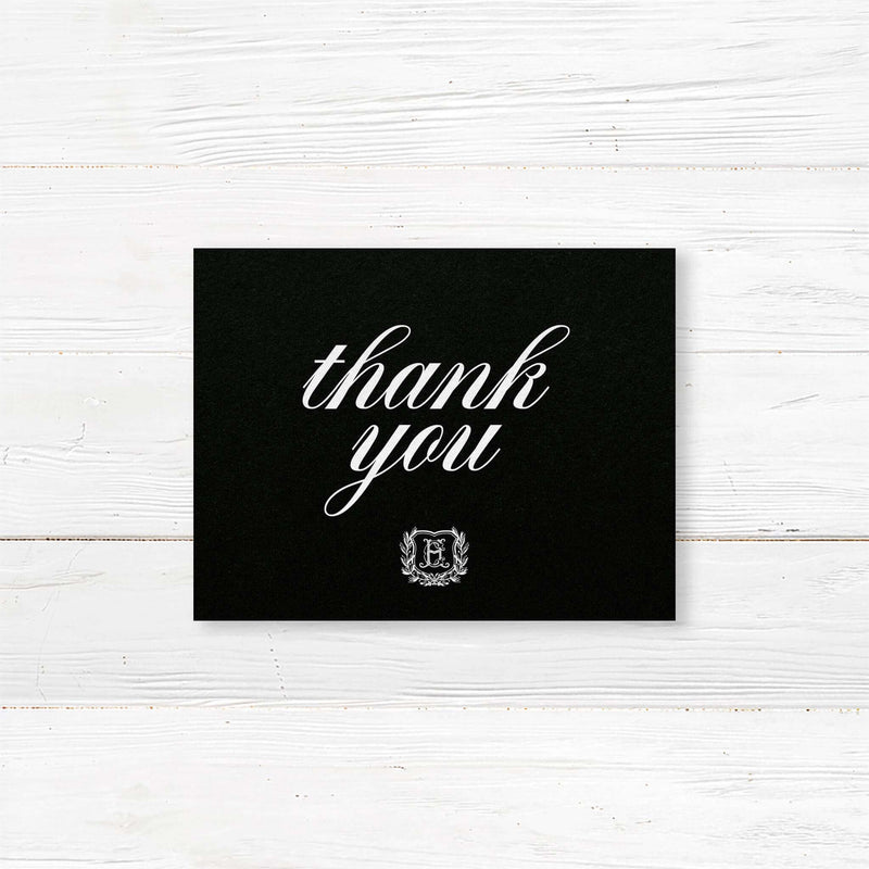 Wedding thank you card with black background, white calligraphy fonts, and an ornate shield monogram encircled by a laurel wreath, matching a formal black wedding suite.