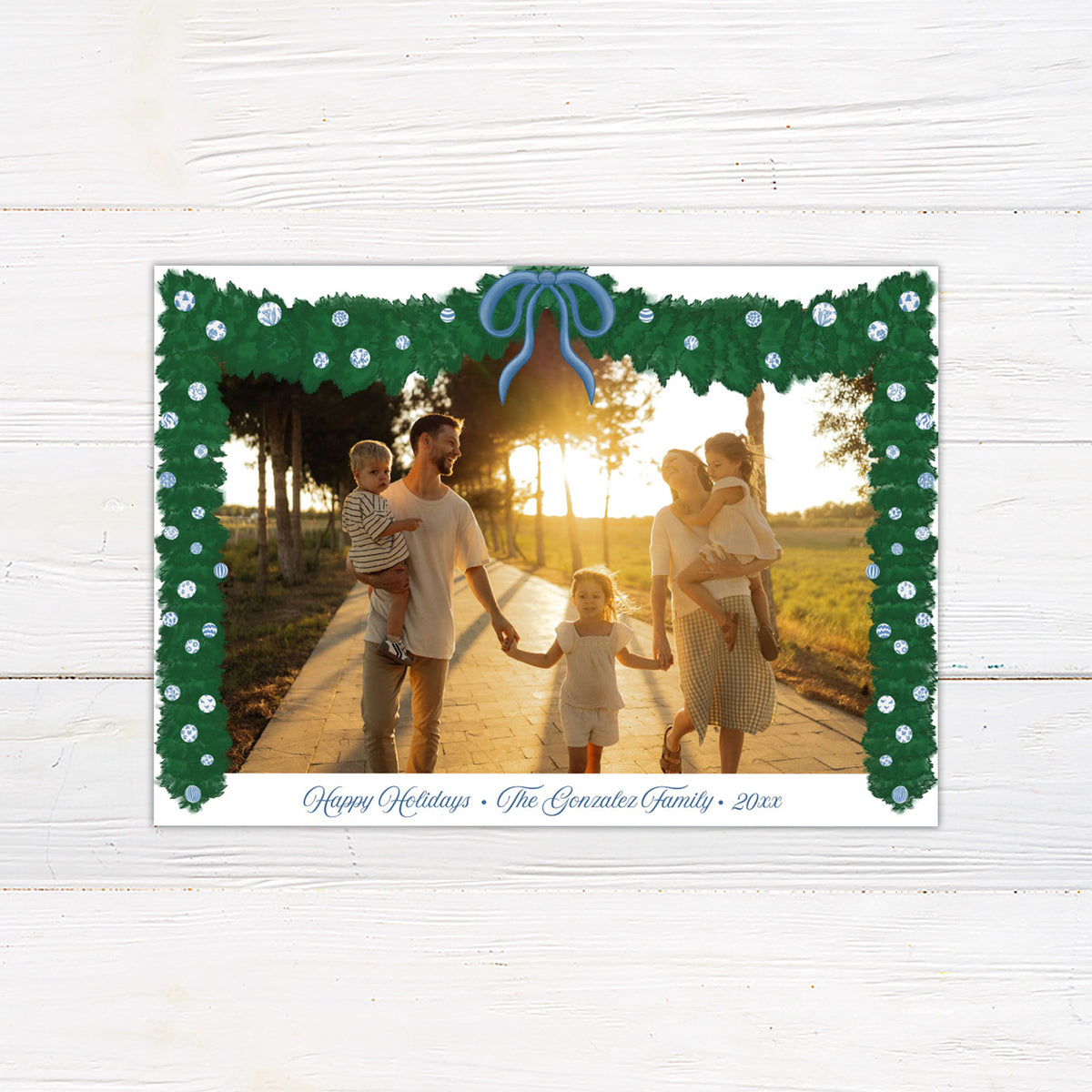 A personalized blue Christmas garland family holiday photo card framed with watercolor evergreen garland, blue and white ornaments, and a blue bow. The coordinating design features elegant blue script and a decorated Christmas tree with matching blue accents. - horizontal front view.