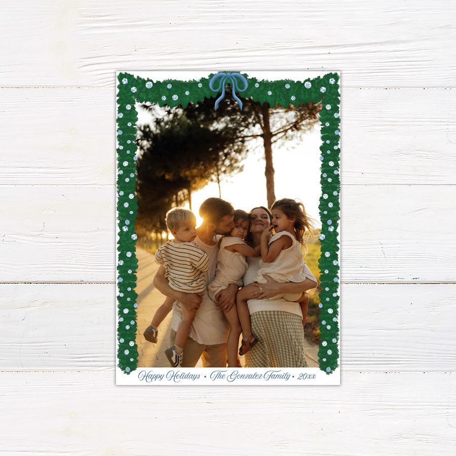 A personalized blue Christmas garland family holiday photo card framed with watercolor evergreen garland, blue and white ornaments, and a blue bow. The coordinating design features elegant blue script and a decorated Christmas tree with matching blue accents. - vertical front view.