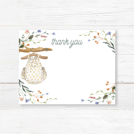 Printed baby shower thank you card featuring a watercolor baby romper hanging on a wooden hanger, surrounded by floral accents. Professionally printed on high-quality cardstock with a matte or glossy finish. Available in flat or folded styles with optional envelopes. Ideal for expressing gratitude after a boho, floral, or rustic baby shower. Complements a gender-neutral baby sprinkle or woodland baby shower theme for a cohesive and elegant stationery set